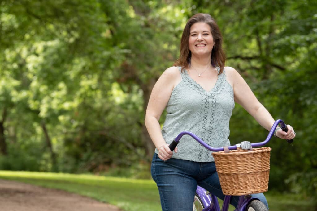 A woman wearing a lacy camisole top and jeans rides a bicycle in a green, wooded environment.