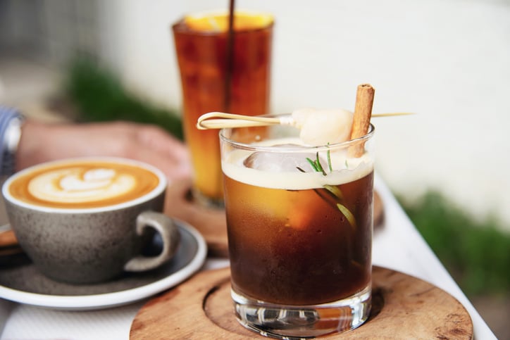 On a table stand a glass of iced tea, a latte and a beverage garnished with a cinnamon stick and rosemary.