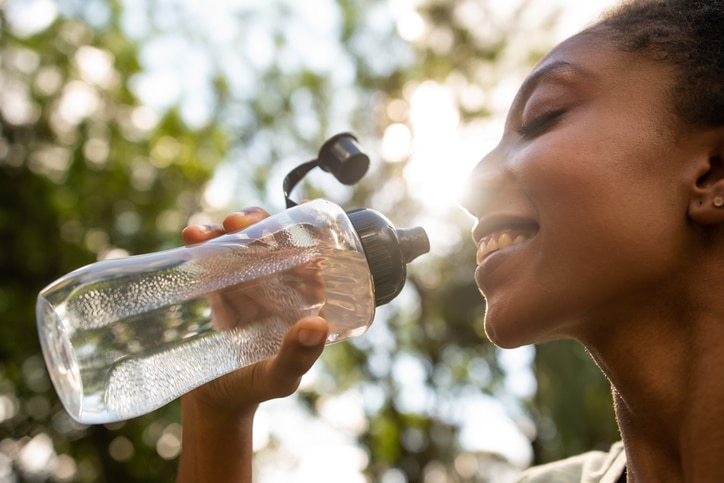 A woman in an outdoor setting drinks water from a clear plastic water bottle.