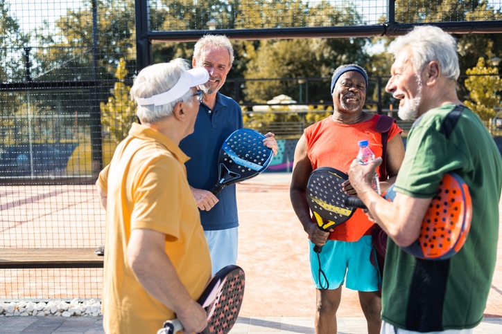 Four older men holding pickleball rackets talk cheerfully against a backdrop of green and gold leaves.