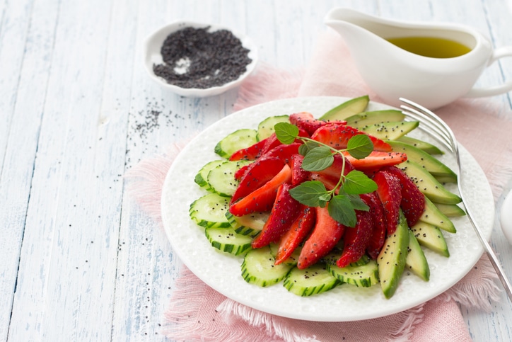 On a white board table, a plate contains cucumber, strawberry and avocado slices. Behind the plate is a small dish of a black spice and a gravy boat containing oil.