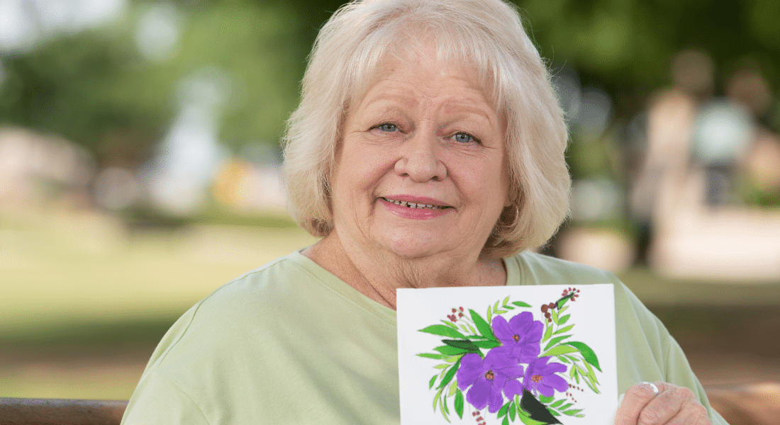 Joan Daniels, photographed and holding a painting she made on a bench after her lung cancer treatment