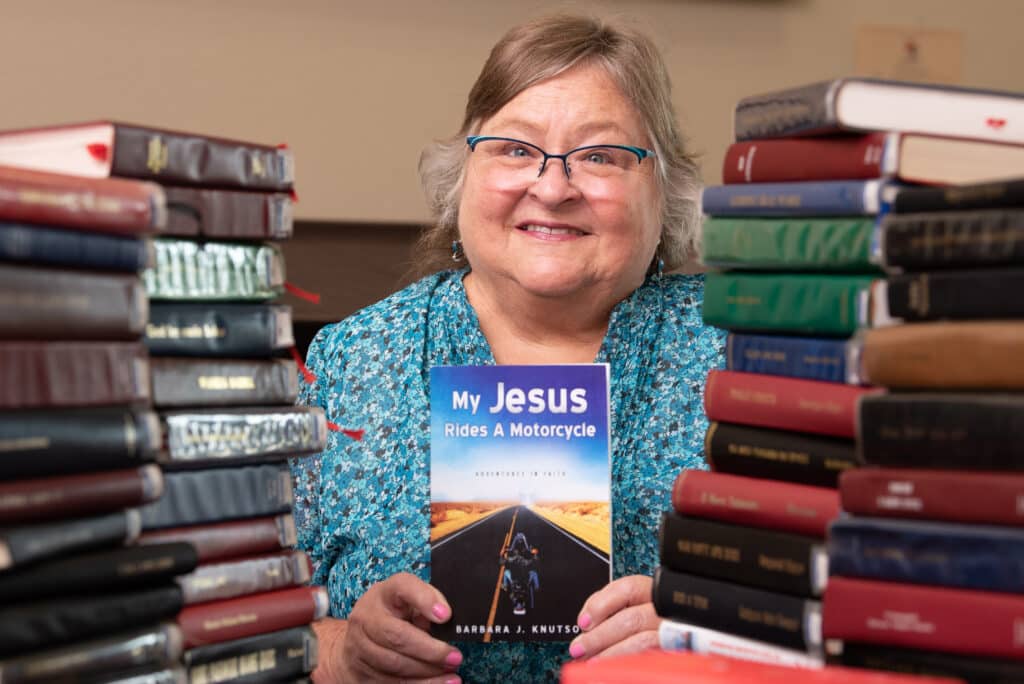 Surrounded by stacks of books, missionary Barbara Knutson shows a copy of her book, My Jesus Rides a Motorcycle.