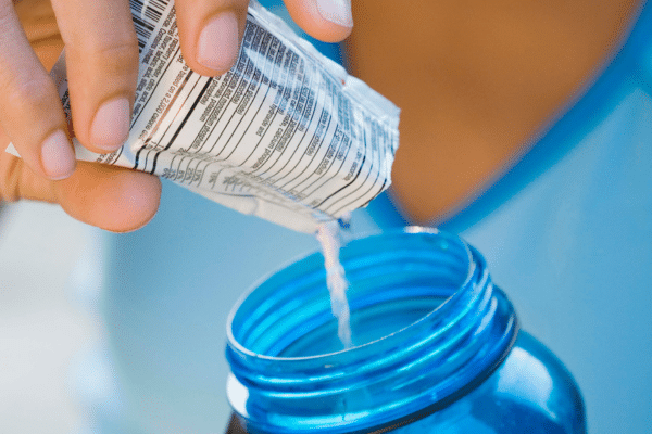 In a close-up shot, a person wearing blue pours a supplement from a packet into a blue plastic water bottle. The nutrition information shows on the side of the packet.