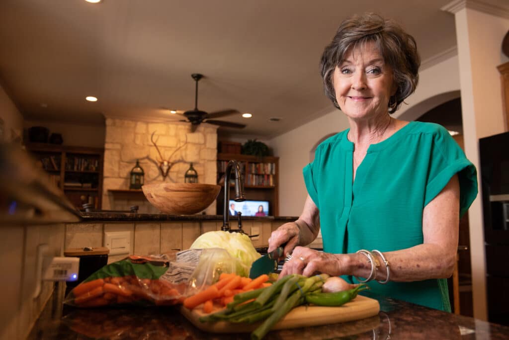 In this photo, a woman chops vegetables, including carrots and cabbage. She is smiling at the viewer.