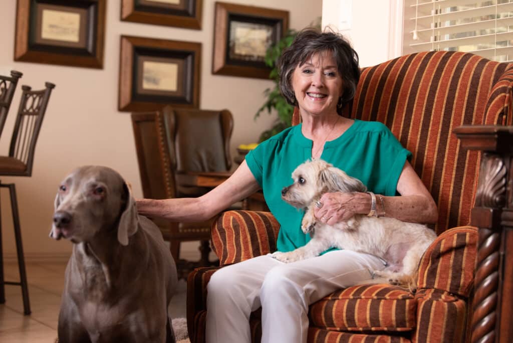 Deborah Sullivan, hip surgery recipient, sits in a striped chair holding a small dog on her lap and petting a large dog beside her. She is smiling happily.