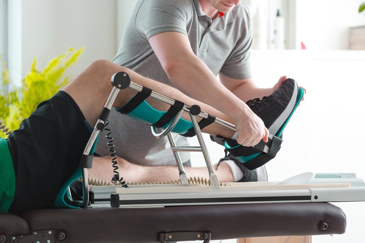 A man wearing gym shorts lies on a table with his leg in a piece of therapeutic equipment. Another man helps adjust the first man's foot into place.