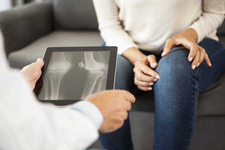 A woman sits in a chair facing the viewer. She is holding her knee. Facing away from the viewer, so that we see over his shoulder, a medical provider holds a tablet showing an X-ray image of a joint.