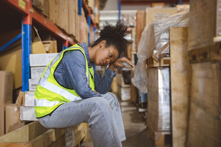 A young woman sits in a warehouse. She wipes her head with the back of her hand.