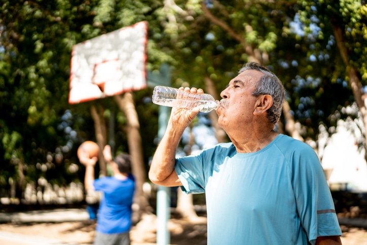 An older man wearing a blue shirt drinks water from a plastic bottle. Behind him, a younger person in a blue shirt plays basketball.