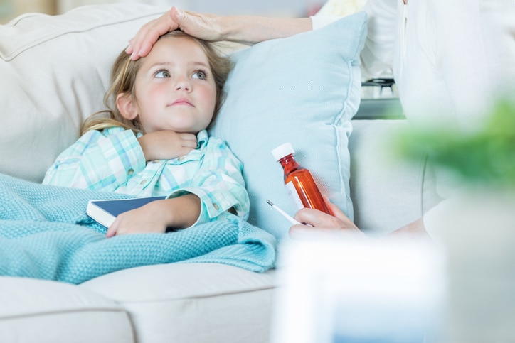 A young child laying on the couch under a blanket while an adult holds their forehead in one hand and medication in the other