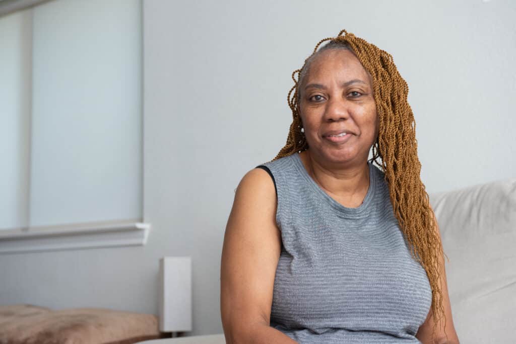 A woman in a gray shirt sits in a room with light-colored walls and furnishings.