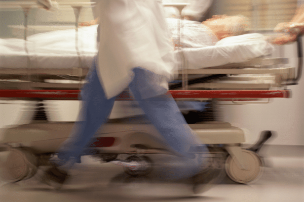 A blurred stock photo of someone in scrubs walking by a stretcher in a hospital hallway