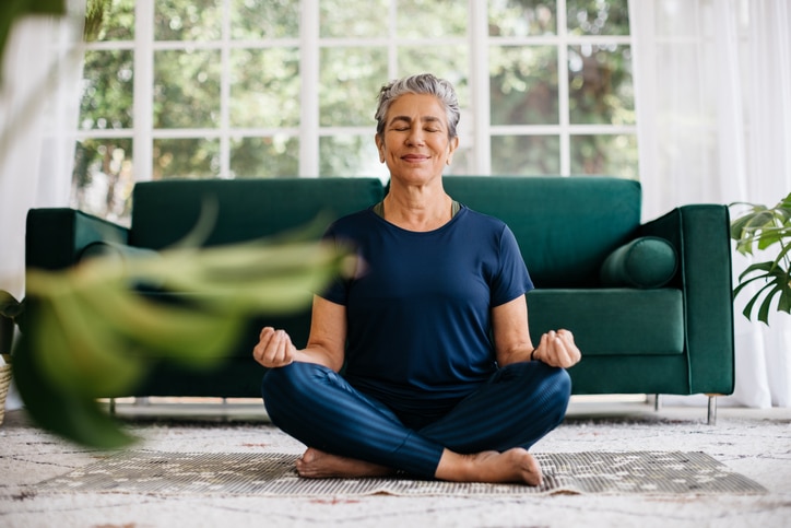 A woman meditates on a rug in front of a green sofa.