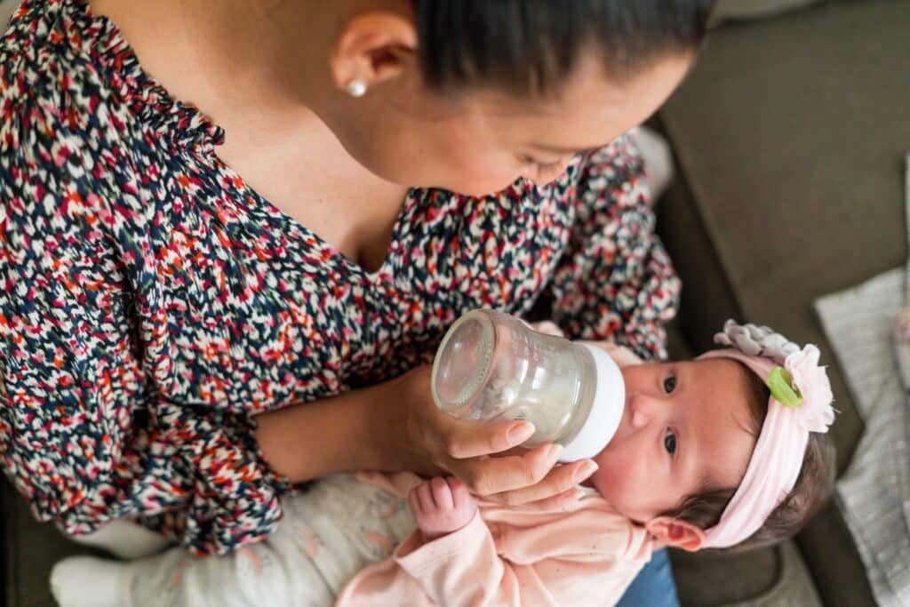 Luz Villagomez, a woman with brown hair pulled back in a bun and a red and black floral blouse, photographed feeding her infant with a bottle