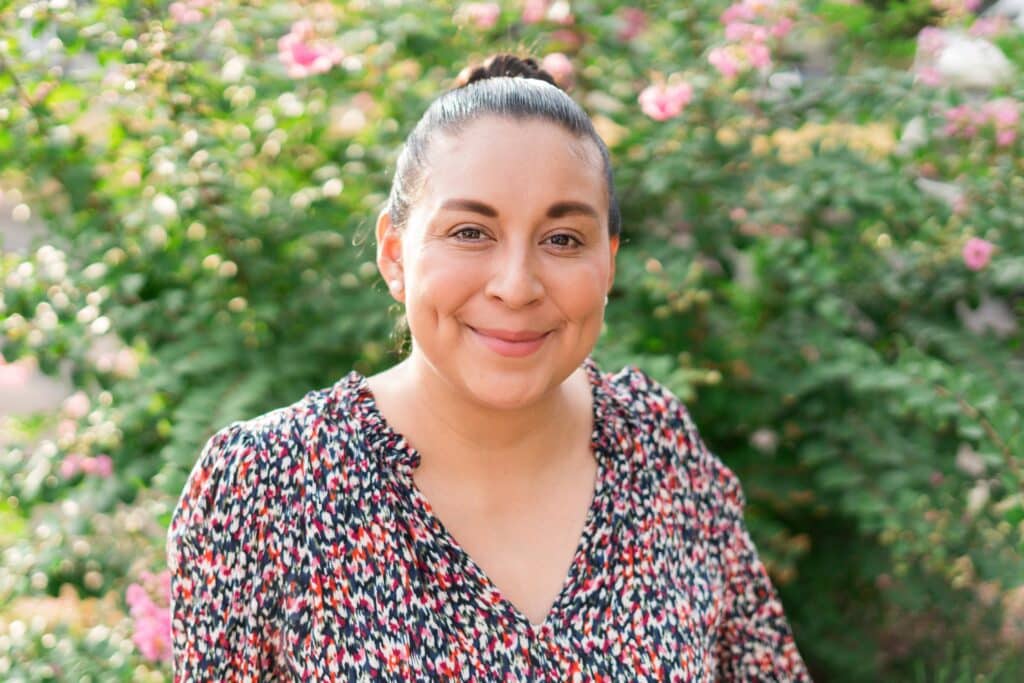 Luz Villagomez, a woman with brown hair pulled back in a bun and a red and black floral blouse, smiles at the camera