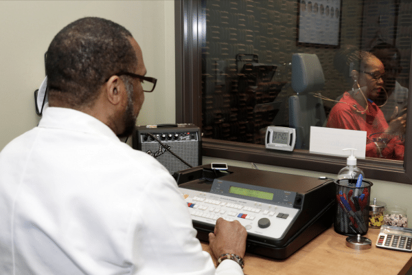 Michael Young conducts a hearing test for a young woman seen through a glass window.