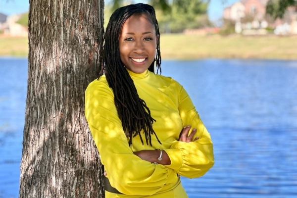 A woman named Thandi Montgomery wearing a bright yellow shirt and smiling and leaning on a tree with a lake in the background