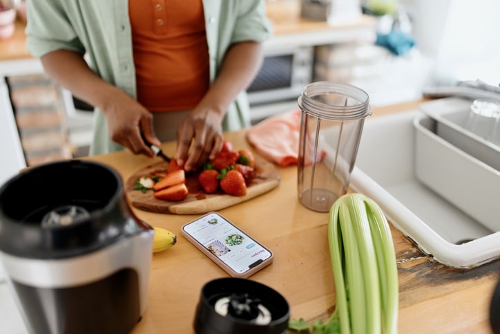 A person chopping fruit and vegetables at a cutting board while looking to instructions on their phone