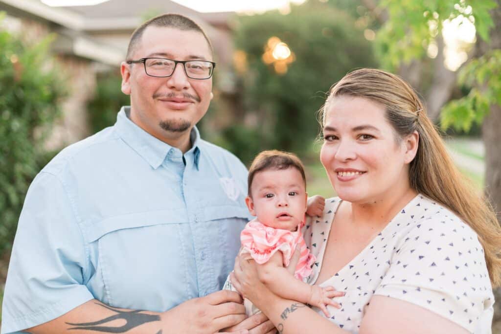 Parents Joe Mendez and Celeste Trevino hold baby Scarlett in an outdoor setting.