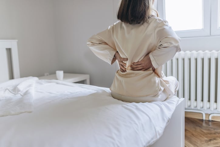 A woman sitting on the edge of a bed stretching her arms behind her back, with her hands grabbing her middle back