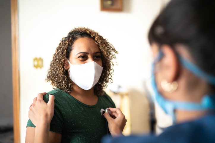 A woman wearing a white face mask having her heart checked by a medical professional with a stethoscope 