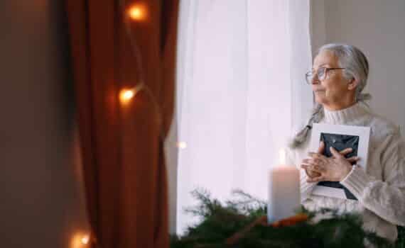 Grieving older woman clutches framed photo in her arms in home with holiday decor.