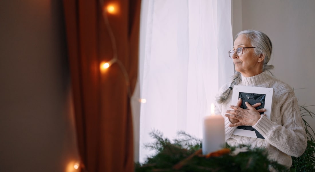 Grieving older woman clutches framed photo in her arms in home with holiday decor.