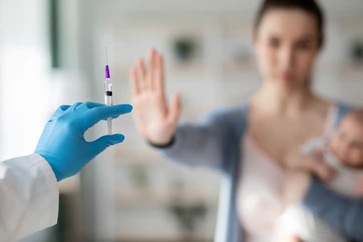 A hand wearing a blue glove holds out a syringe. In the background, a woman holding a baby lifts her palm in a gesture of refusal.