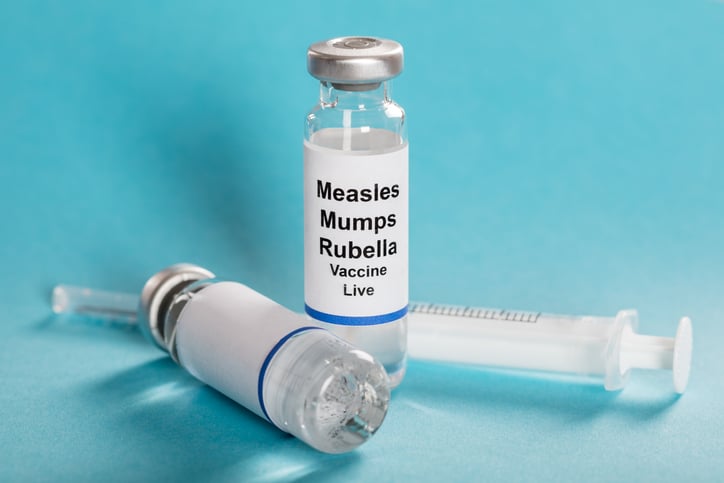A bottle labeled "Measles Mumps Rubella Vaccine Live" stands against a blue background. There is a syringe behind the bottle.