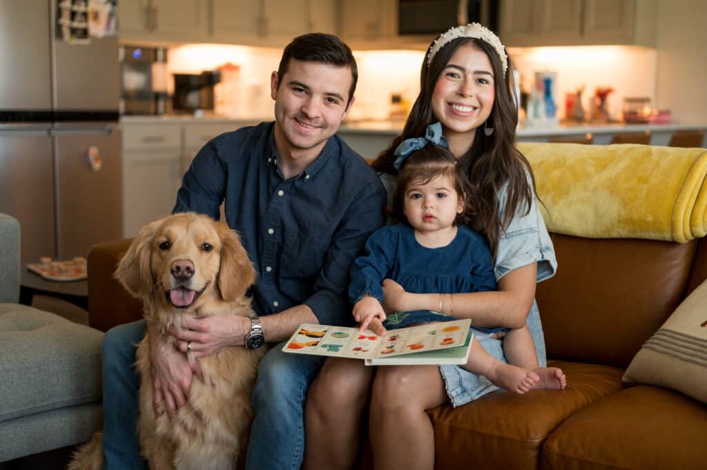 Alexandria Hurst, her husband Micah, and baby Aniston sitting on a couch and smiling for the camera with their dog