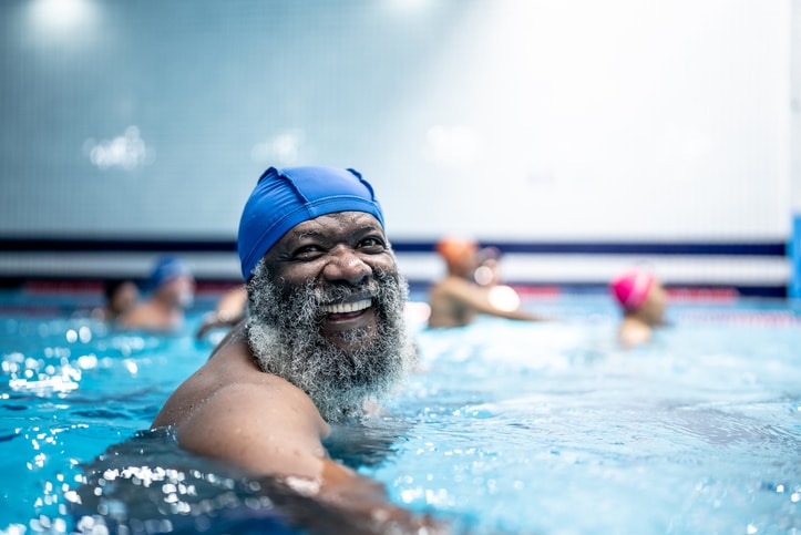 An older Black man with a beard wearing a blue swimming cap while in a swimming pool and smiling at the camera