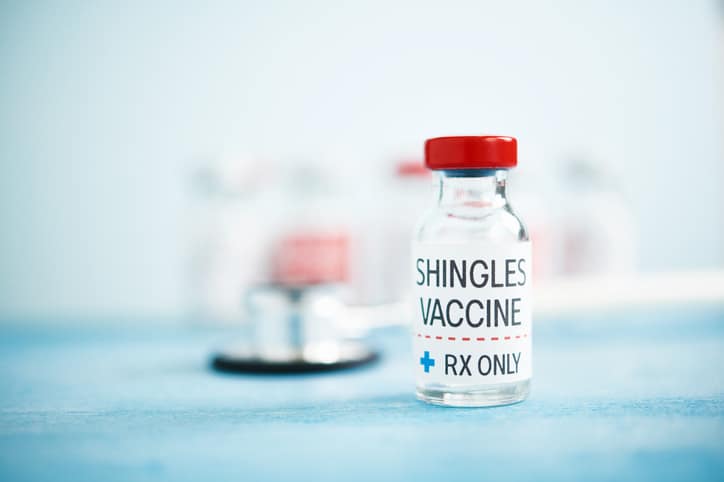 A stock image of a blurred background and a red-capped container with a label that says "SHINGLES VACCINE / RX ONLY" in the foreground.
