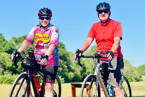 Ed Ofria photographed in a red shirt and black activewear shorts while riding his bike, standing next to another man in a pink riding shirt also riding a bike