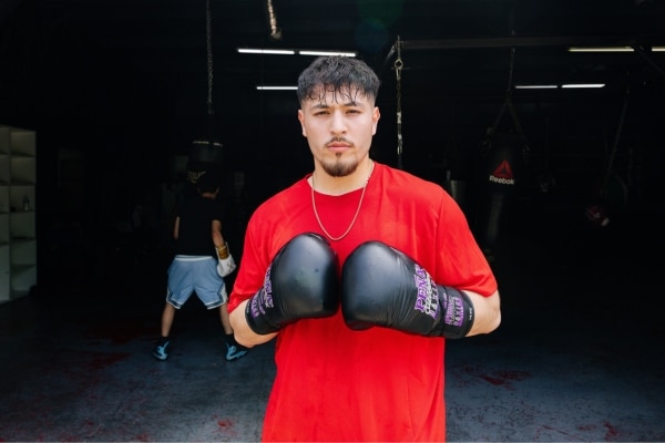 A young man wearing a red shirt and black boxing gloves faces the camera with confidence.