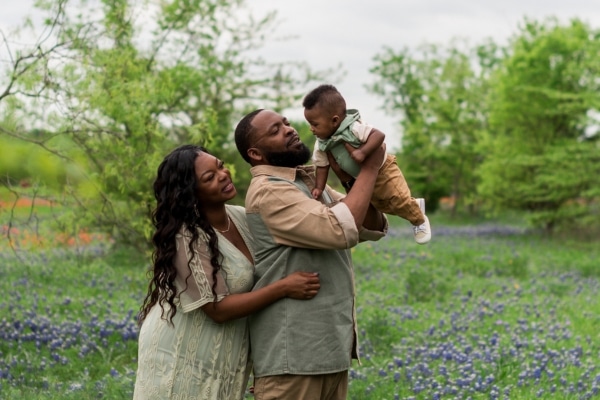 LaTeisha Young, her husband Jeromey, and their newborn Mason photographed outside in an editorial or portrait style while wearing green and brown complementing outfits, looking up at Mason while Jeromey holds him in the air with both hands