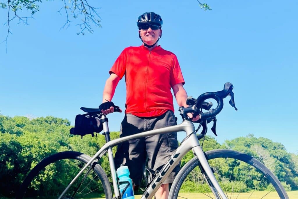 Ed Ofria photographed smiling at the camera while wearing a red shirt and black activewear shorts while standing next to his bike