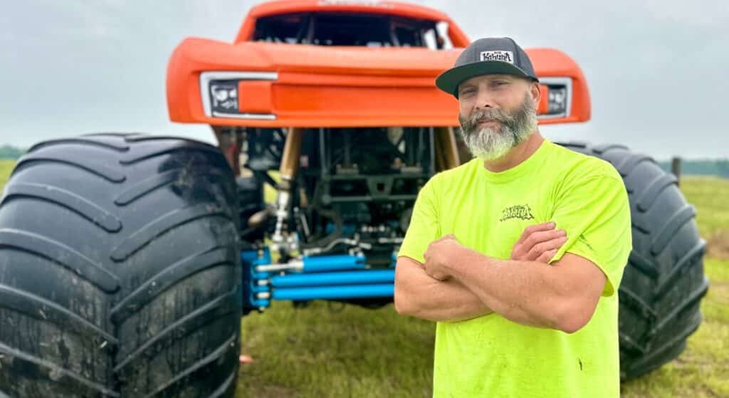 A man named Shane England standing in front of an orange monster truck, used to explain his recovery after a widow maker heart attack