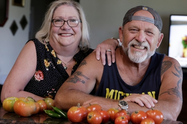 A man and woman smile at the camera. The woman's arm rests on the man's shoulder. In front of them are tomatoes and peppers.