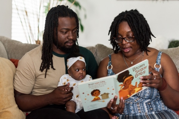 Sitting on a sofa, a man, woman and baby girl look at a book together. The book's cover reads "Brown Sugar Baby."