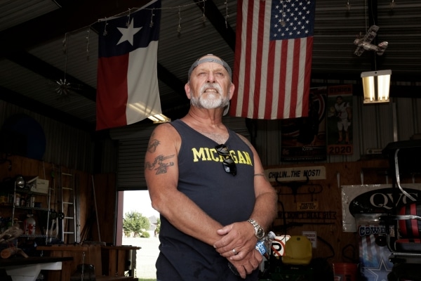 A man wearing a baseball cap and a Michigan athletic shirt poses in a dark interior. An American flag hangs in the background. A door behind him reveals a sunny field.