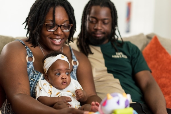 A woman sits on a sofa and entertains a baby girl with a toy. The woman is smiling. Beside her, a man looks happily at the baby.