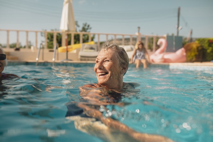 An older woman treads water in a swimming pool on a clear day. She is smiling joyfully.