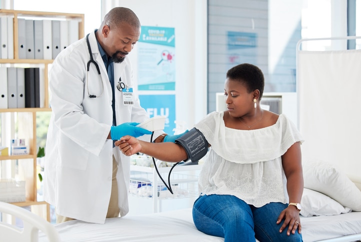 A woman sits on an exam table wearing a blood pressure cuff. A man wearing a white coat and stethoscope is checking her readings. 