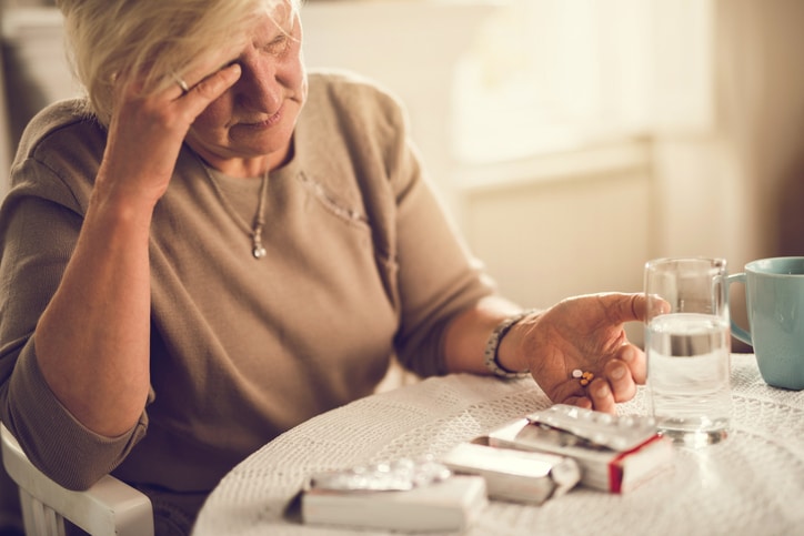 An older woman wearing brown shirt looks at several pill containers in front of her. She holds a few different-colored pills. She's resting her head on her other hand.