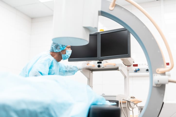 A medical professional in scrubs looks at a dual monitor. In the foreground is a C-arm imaging system.