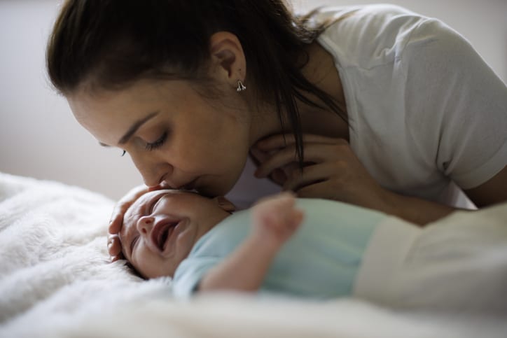 A woman kisses a crying baby who is lying on a fleecy rug.