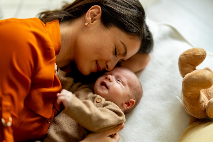 A woman and infant snuggle on a pillow. A teddy bear lies nearby.