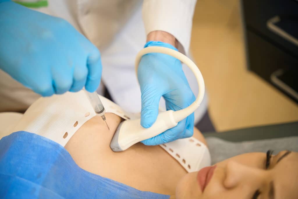 A woman lies on a table. A doctor wearing blue gloves uses an ultrasound detector and a needle to perform a breast biopsy.