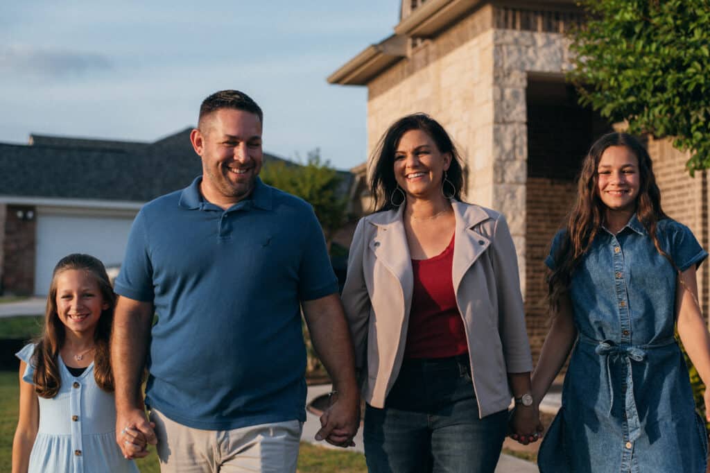 A man, woman, and two girls hold hands in front of a cream-colored brick building.